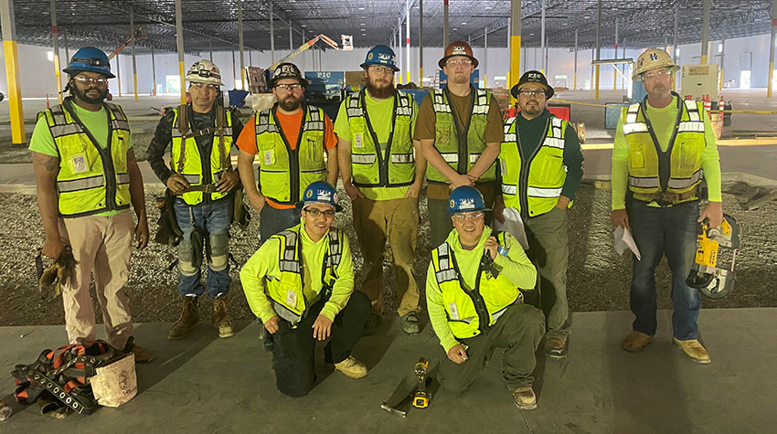 Picture of seven PIC employees in hard hats and safety vests, with two in front holding a safety vest with the PIC logo